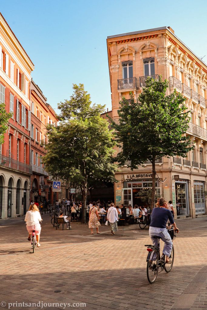 two bikers on the stone streets of toulouse with pink terracotta buildings and a busy patio in the background