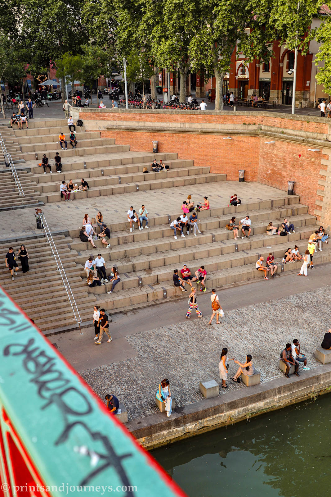 an aerial view taken from the bridge of small groups of people sitting on the stairs lining the river near pont saint pierre