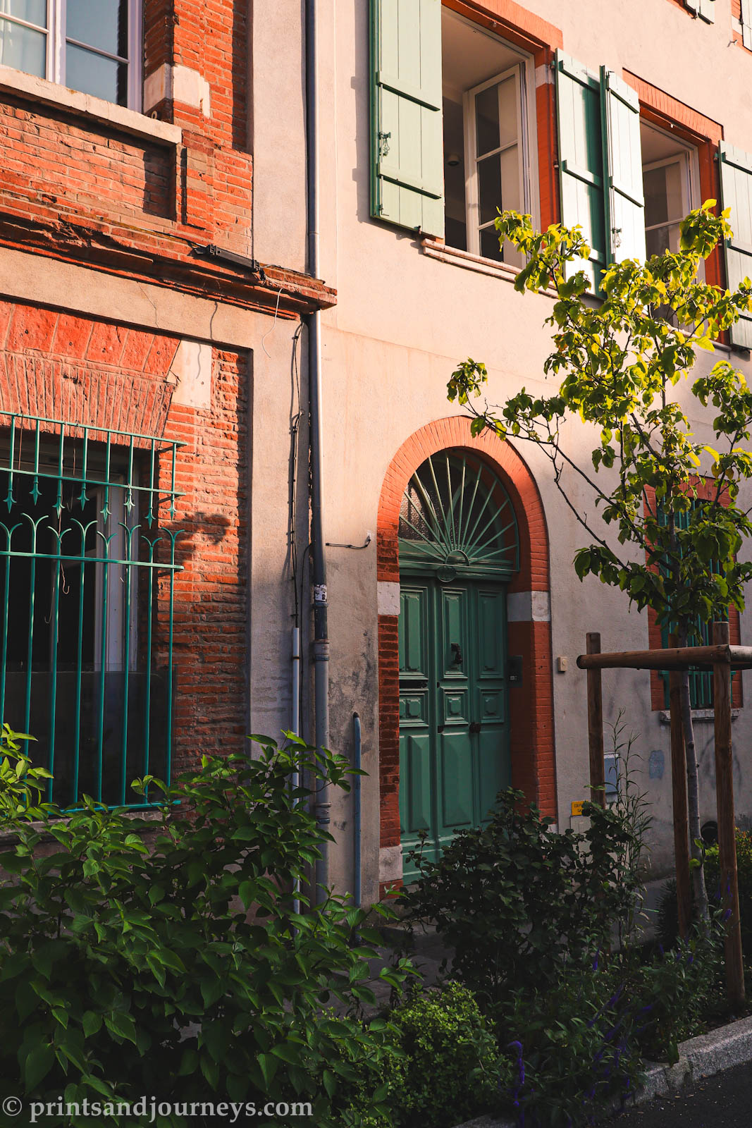 a pink terracotta building in toulouse with the french shutters open letting in the sun and shaded trees in the foreground