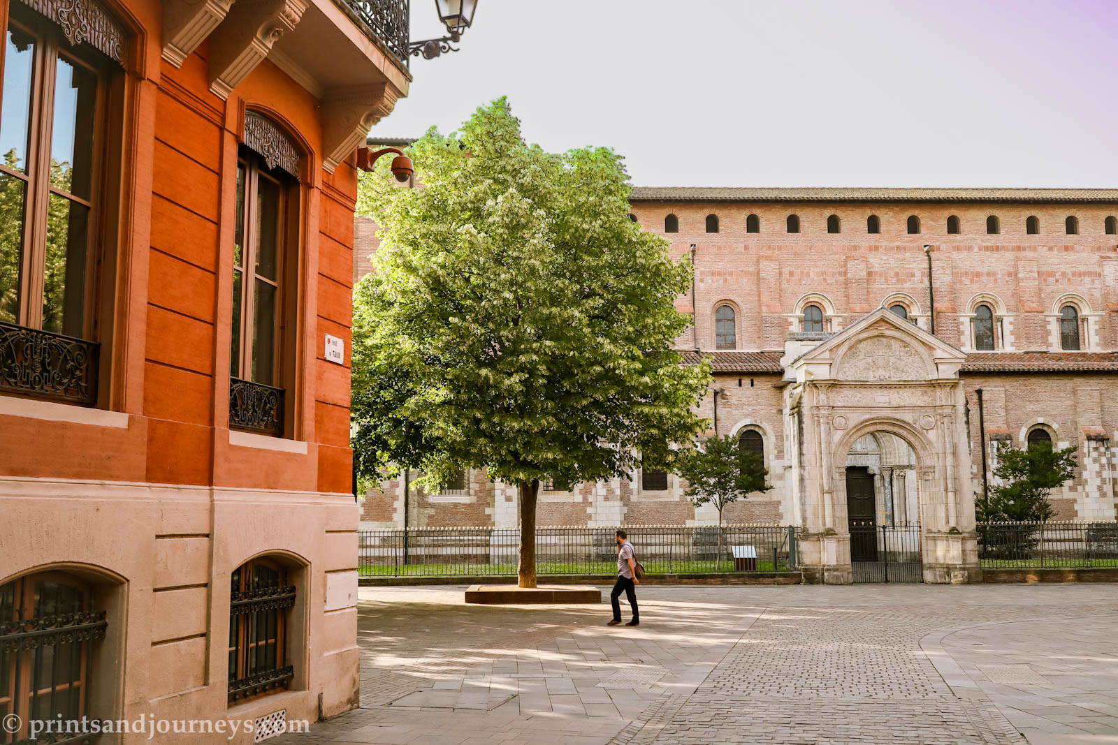 one person walking in a square in Toulouse with red and pink buildings and a green leafy tree